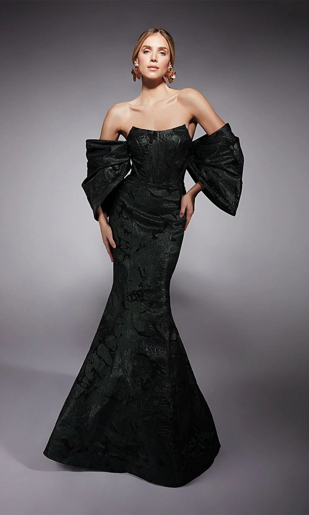 A woman poses in the Alyce 27747 long formal dress, featuring forest green metallic jacquard fabric, off-the-shoulder bow-like sleeves, and a fitted mermaid silhouette, set against a neutral background.